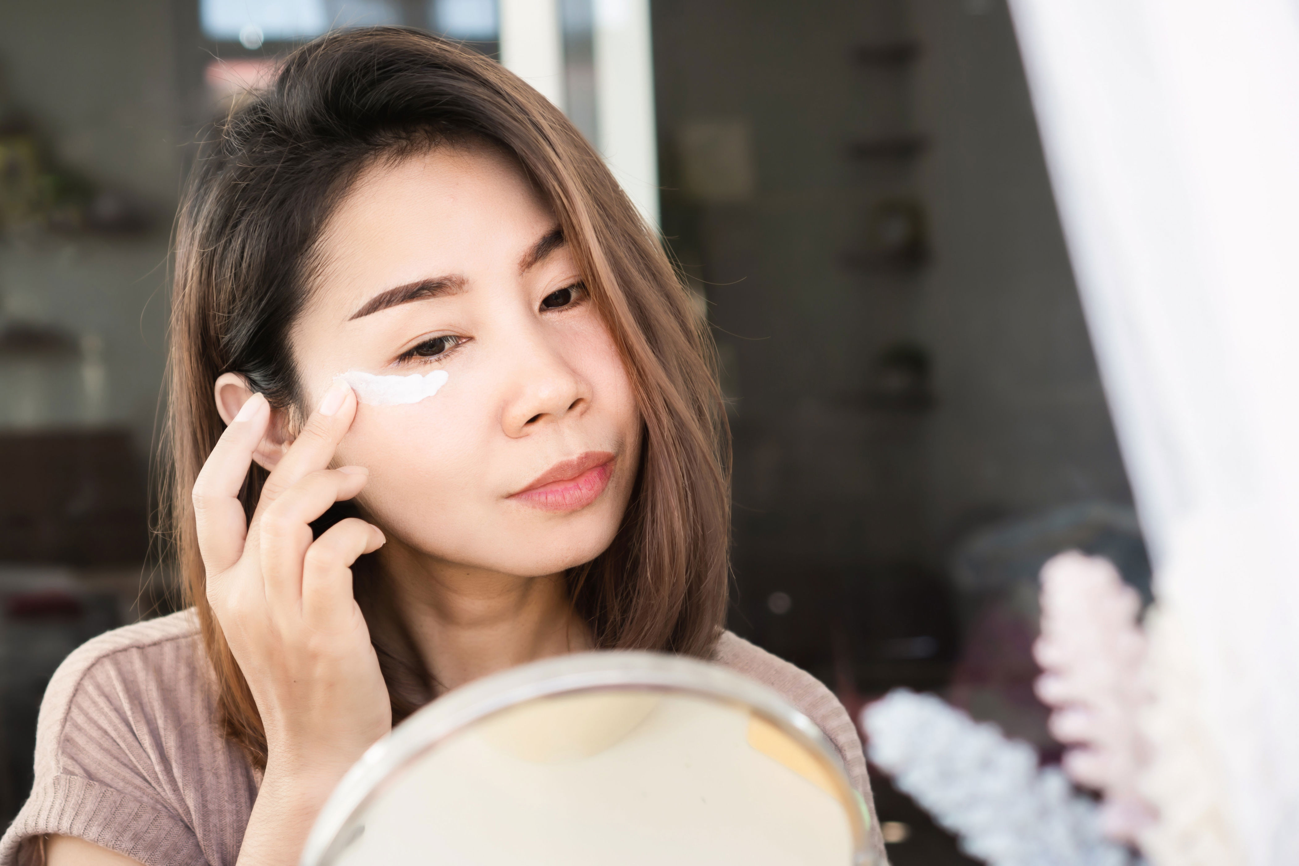 Woman applying face cream while looking in a mirror, focused on skincare routine