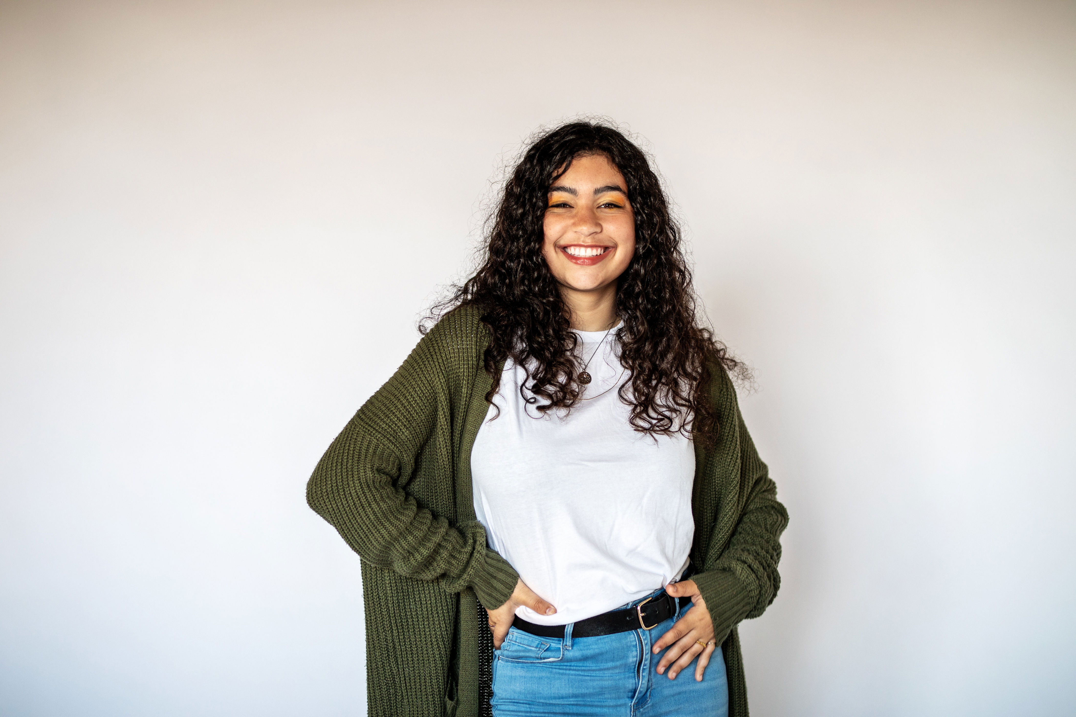 Person smiling, wearing a white shirt, jeans, and a cardigan. Hands on hips, standing against a plain background