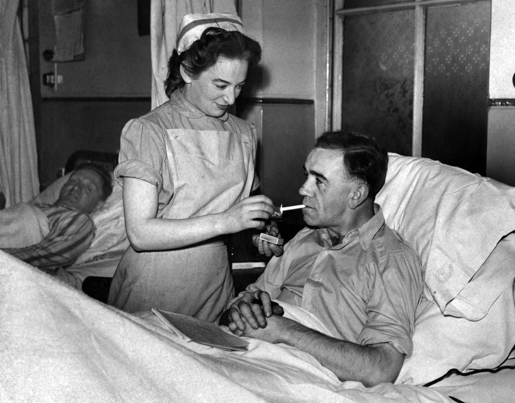 A nurse feeds a male patient in a hospital bed, another patient rests in the background. Mid-20th century healthcare setting