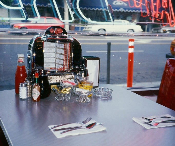 Retro diner table with a jukebox, condiments, and folded napkins. Neon lights reflected in the window