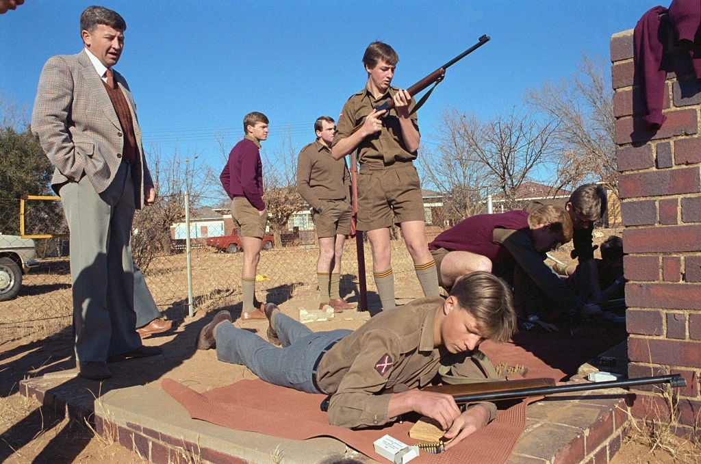 A group of people in uniform participate in a rifle training exercise outdoors, supervised by an adult