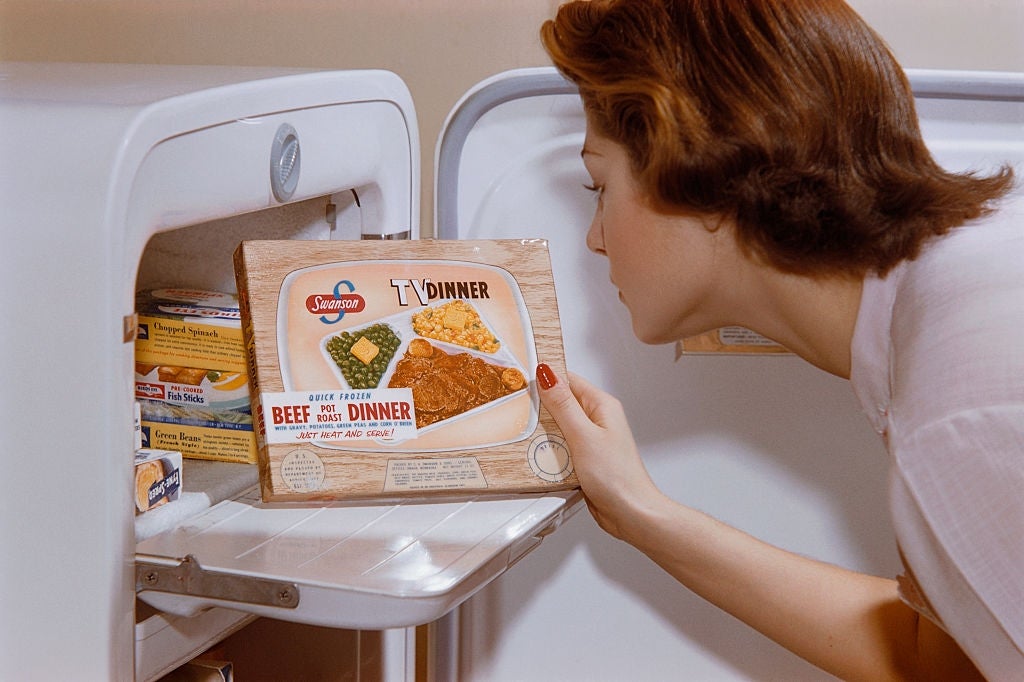 A woman in 1950s style looks into a freezer, holding a TV dinner box with beef, peas, and a pie, symbolizing mid-century convenience food