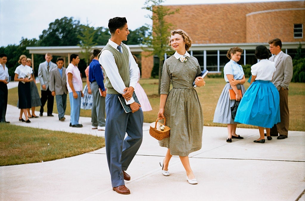 1950s students chat on a school campus. A young man in a vest walks with a young woman in a checked dress carrying a book and a basket