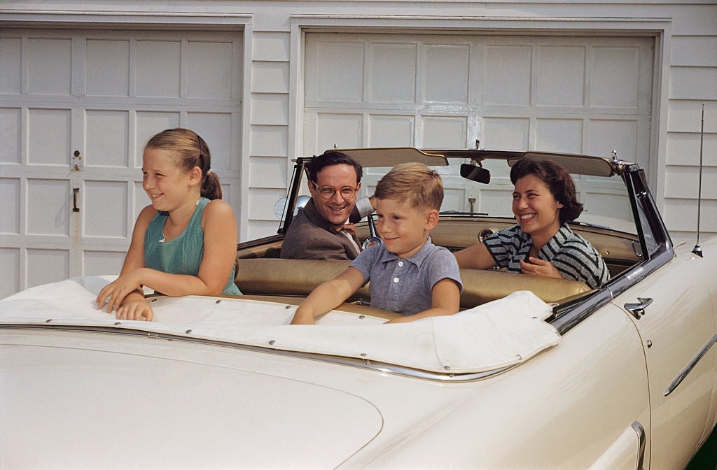 A family of four smiles while sitting in a vintage convertible parked in a driveway, with the top down