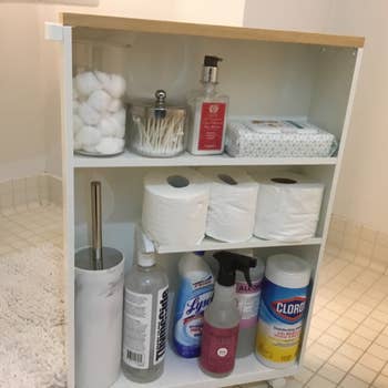 Bathroom storage cart with cotton balls, swabs, tissues, hand soap, toilet paper, a plunger, and various cleaning supplies