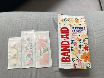 Box of Band-Aid flexible fabric bandages with floral patterns, shown with three open bandages displaying different flower designs