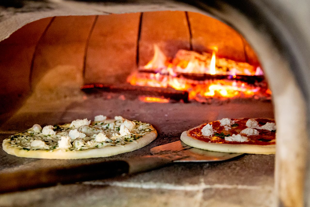 Two pizzas being baked inside a woodfired oven with visible flames in the background