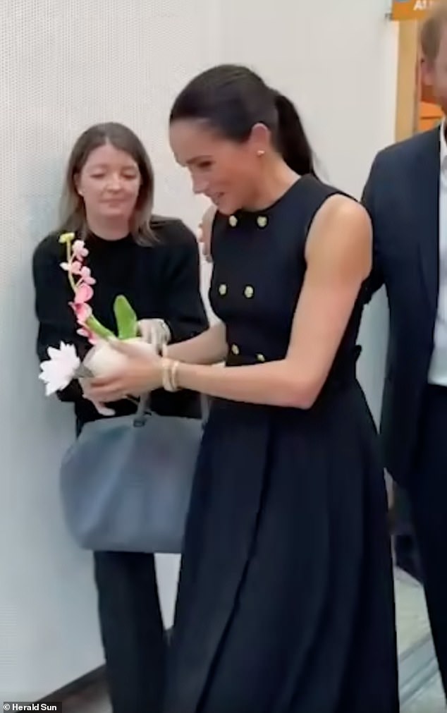 Just like a working royal! Moment Meghan hands flowers to aide during ...