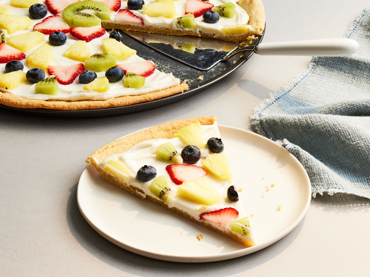 Close up on a slice of fruit pizza on a plate with remaining on pizza tray behind.