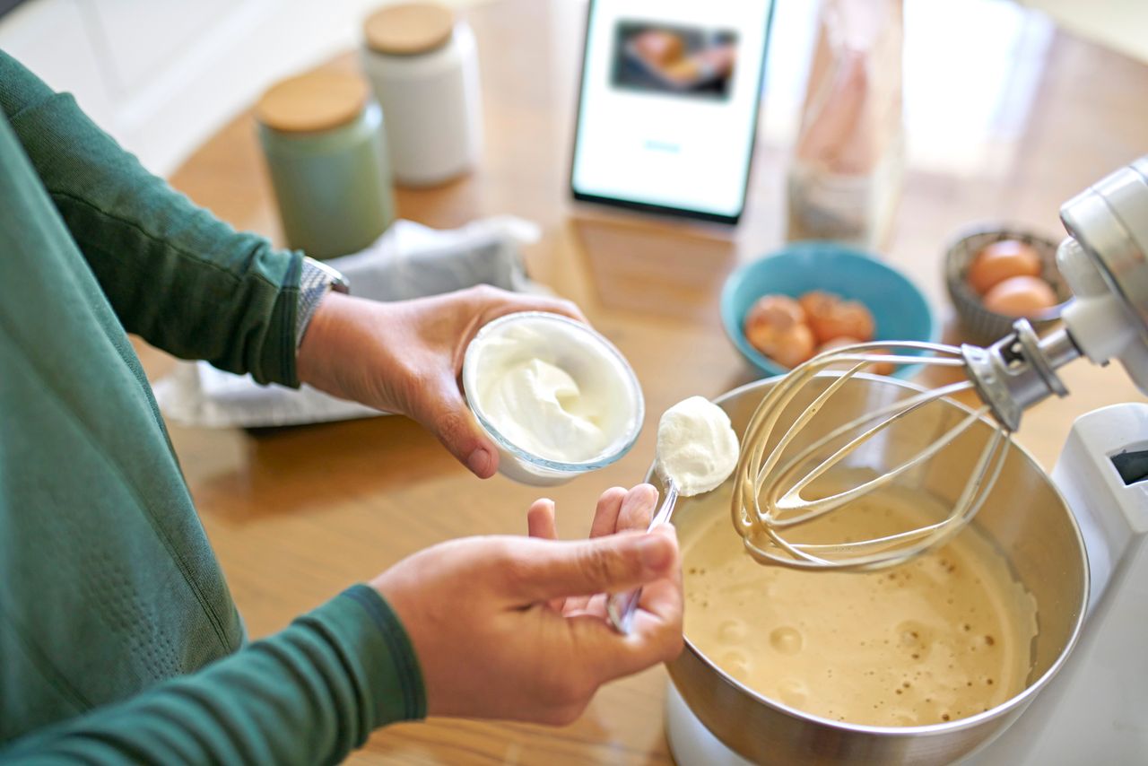 A person adding sour cream to a bowl of batter in a stand mixer with baking ingredients on the table in the background