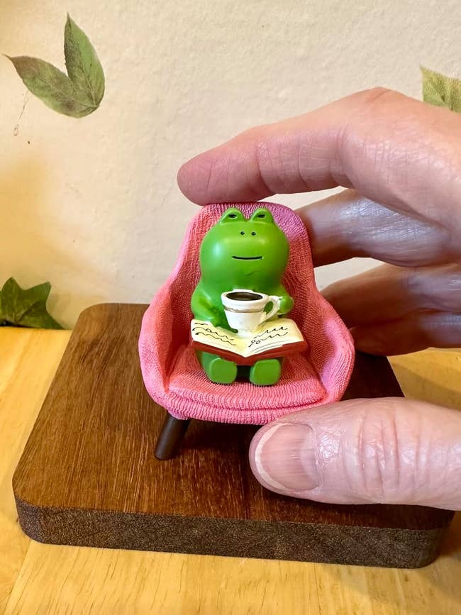 Figurine of a frog sitting in a fabric chair, holding a book and cup; someone's hand nearby. Suitable for home decor