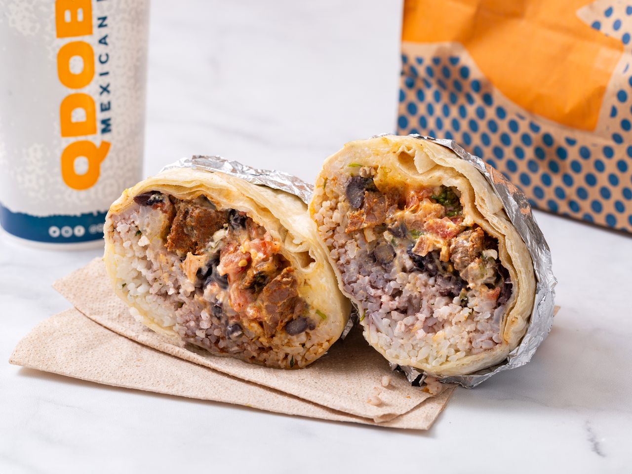 A grilled steak burrito from Qdoba, cut in half, with a drink and bag in the background