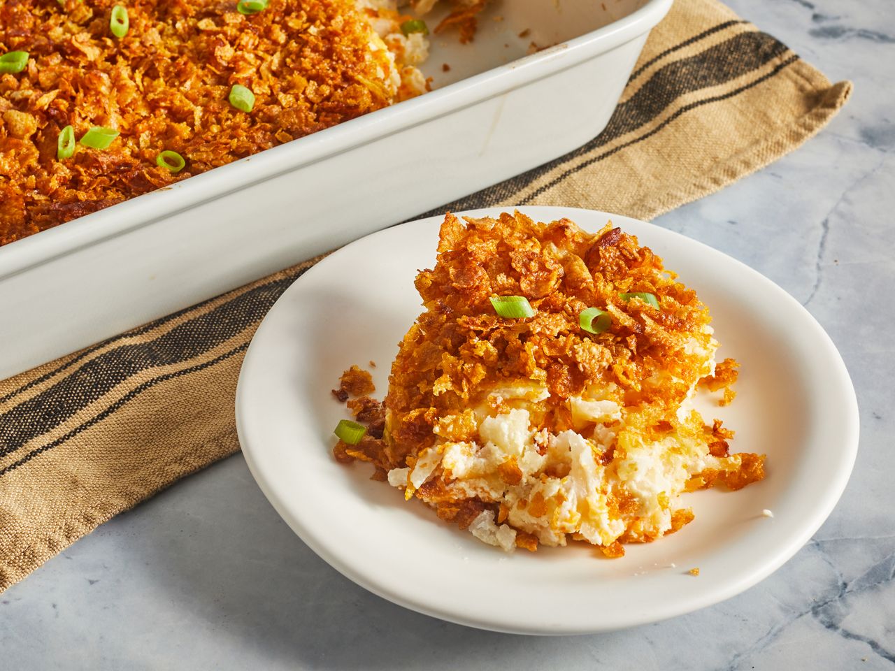 a low angle, close-up view of a heaping scoop of cheesy potato casserole plated in front of a casserole dish with remaining potato casserole garnished with green onions