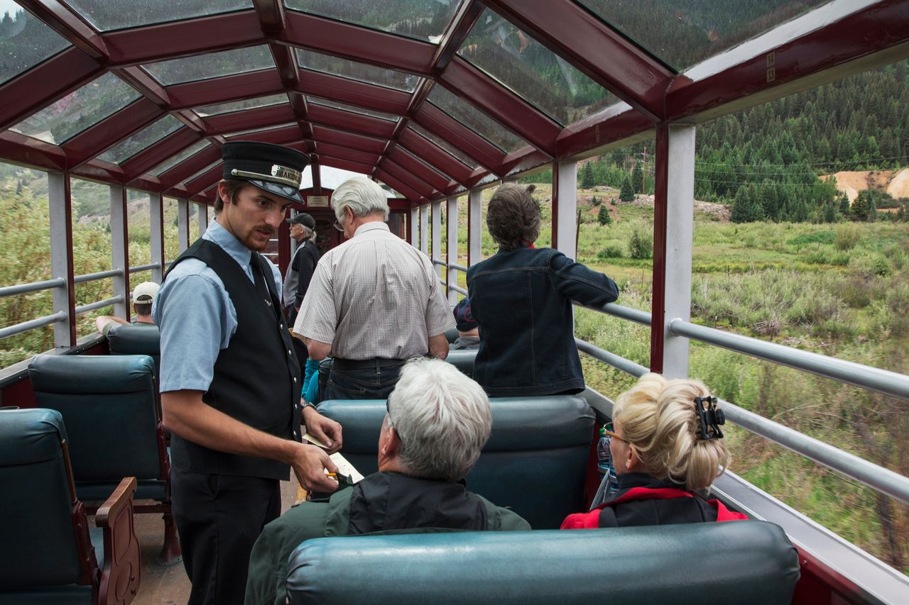 Train conductor interacting with passengers on an open viewing car of a train with scenic rural landscape visible outside