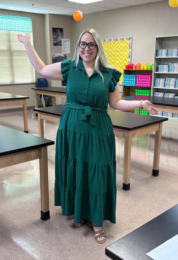 Person standing in a classroom with wooden desks, wearing a tiered dress and glasses, smiling and gesturing welcomingly
