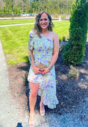 Person outdoors in a floral, asymmetrical dress, holding a clutch, smiling on a sunny day with greenery and trees in the background