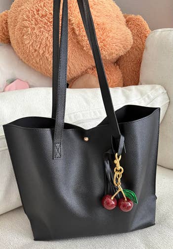 Black tote bag with cherry charm on sofa; large teddy bear and cushion backdrop
