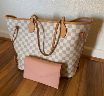 Checkered tote bag with brown handles and a folded pink clutch placed in front, set on a wooden floor