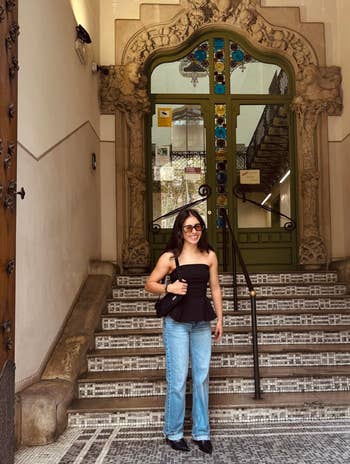 A person stands on a tiled staircase, wearing sunglasses, a strapless top, jeans, and holding a bag, in front of a decorative entrance