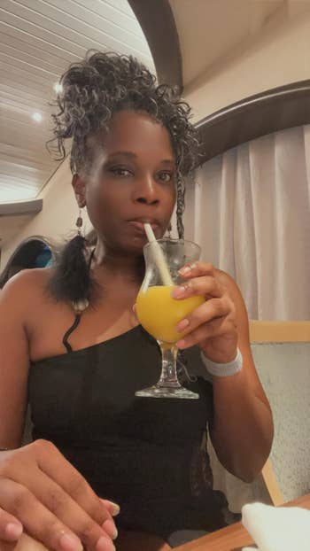 Person in an elegant black outfit drinking a yellow beverage from a fancy glass at a dining setting
