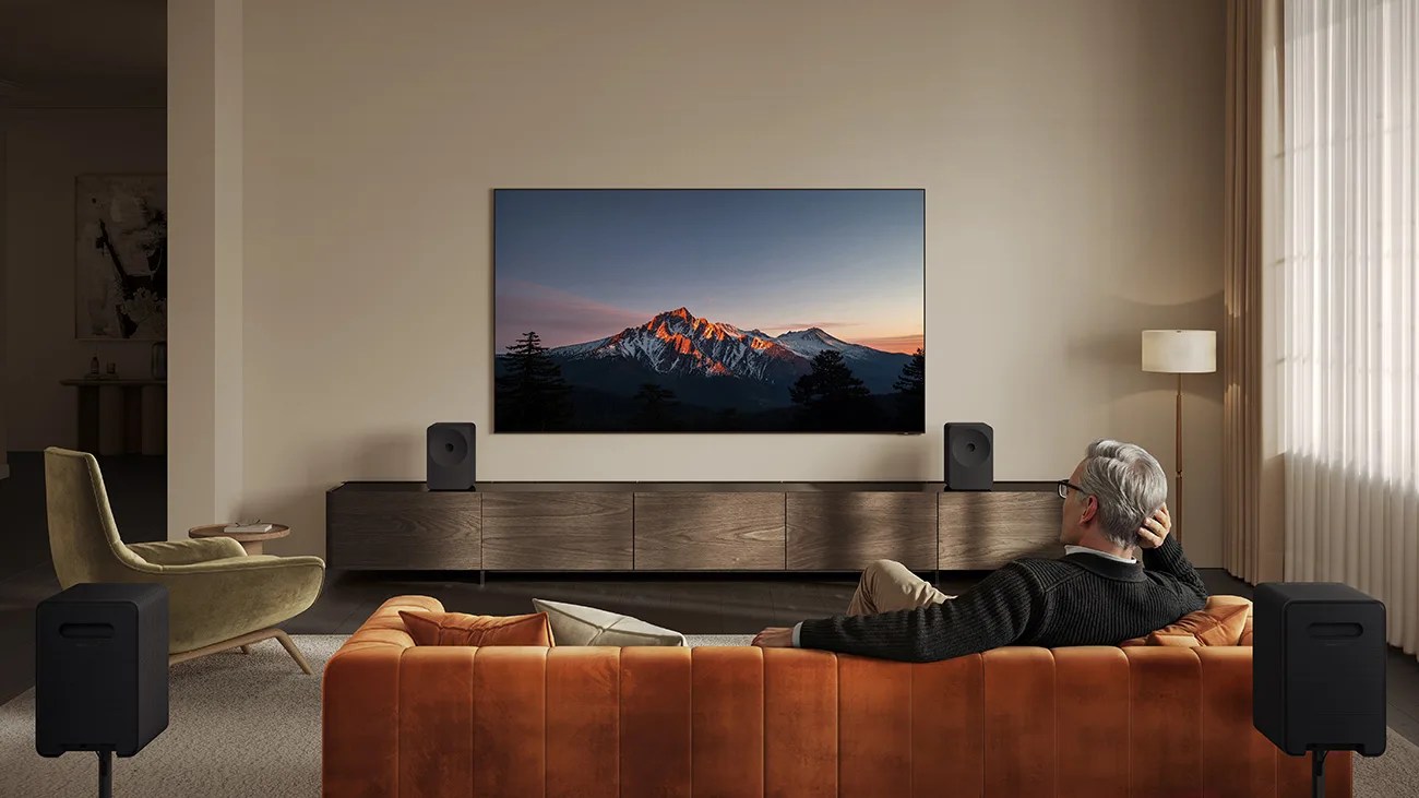 Modern living room with a wall-mounted TV showing a mountain scene, brown leather sofa, and black speakers.
