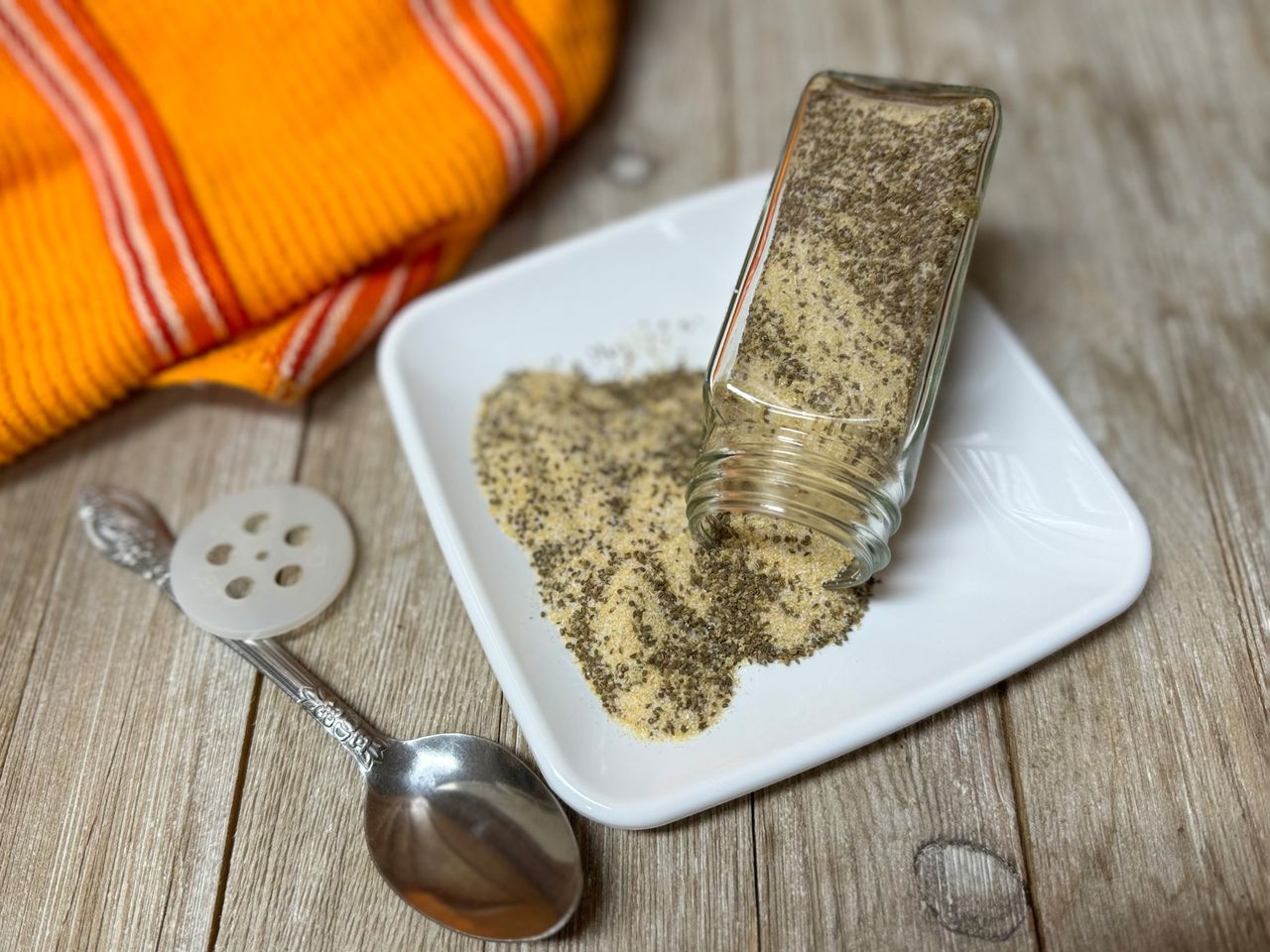 spice jar with beau monde seasoning mix pouring onto small white plate, spoon and orange towel in background
