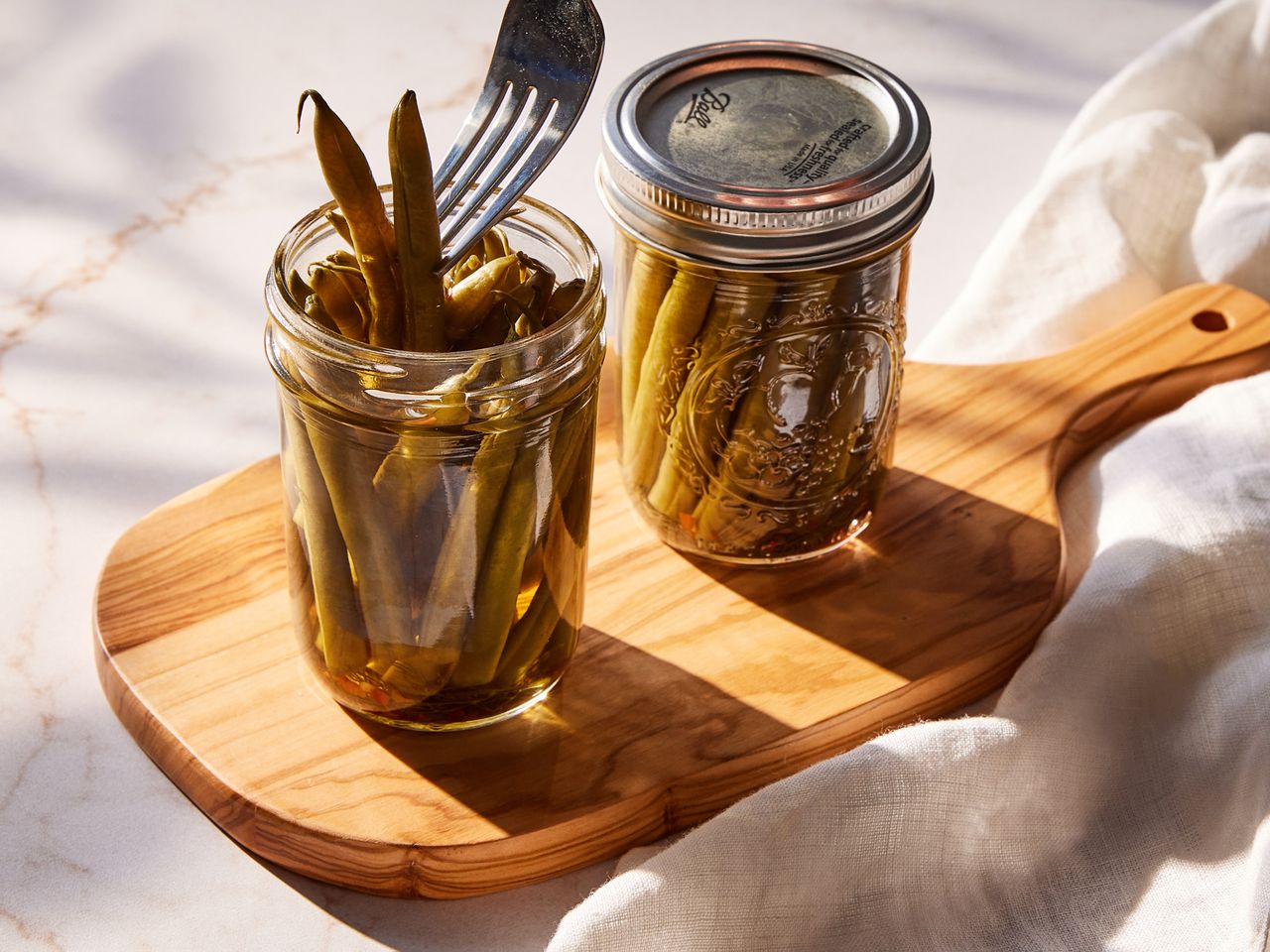 Two jars of crisp pickled green beans on a cutting board, with a fork removing two green beans from an open jar