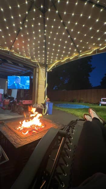 Person relaxing by a fire pit under string lights, watching a movie projected on a screen in a backyard setting