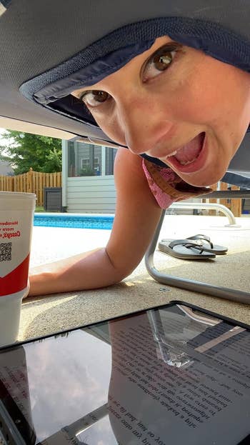 Person grinning under a sunshade near a pool, with a tablet displaying text and a cup nearby