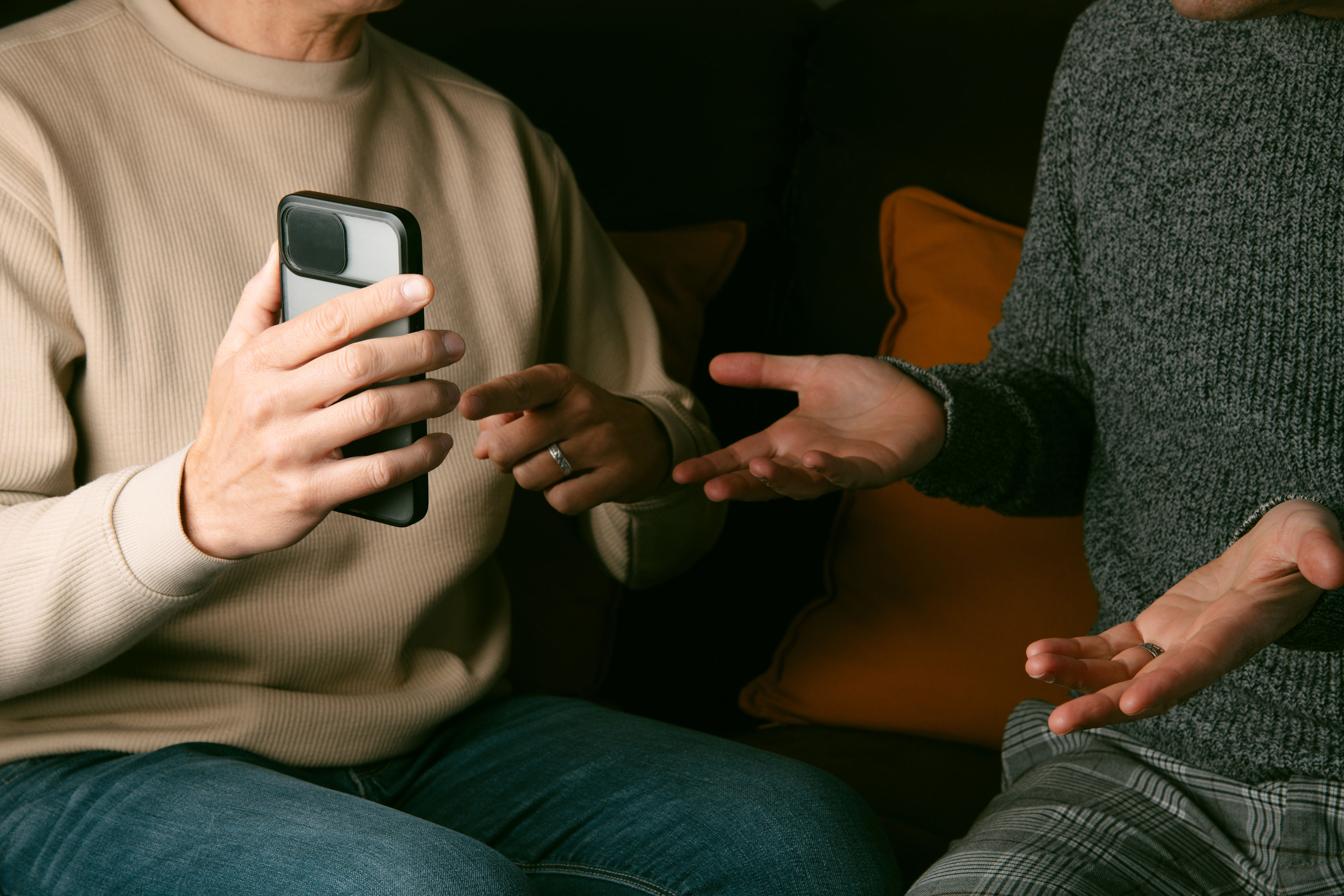 Two people sitting on a sofa, one holding a smartphone, engaged in a conversation with expressive hand gestures