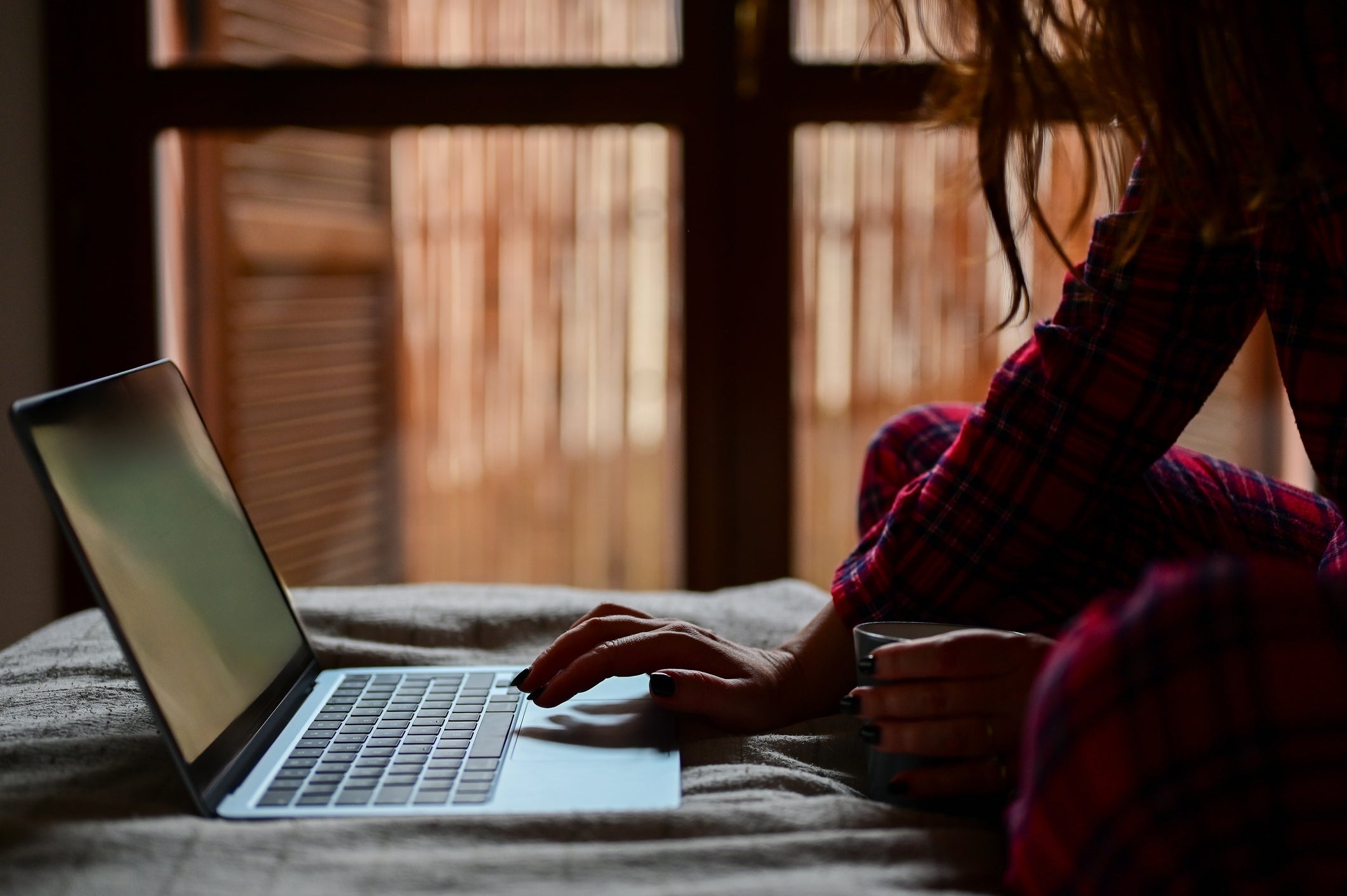 Person in pajamas using a laptop on a bed, holding a mug, in a cozy dimly-lit room