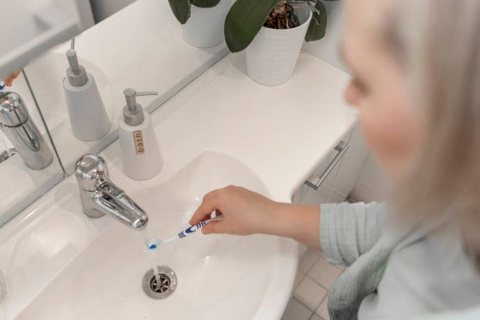 Person cleaning a bathroom sink with a toothbrush, focusing on the drain area. Soap dispensers and a potted plant are visible on the counter
