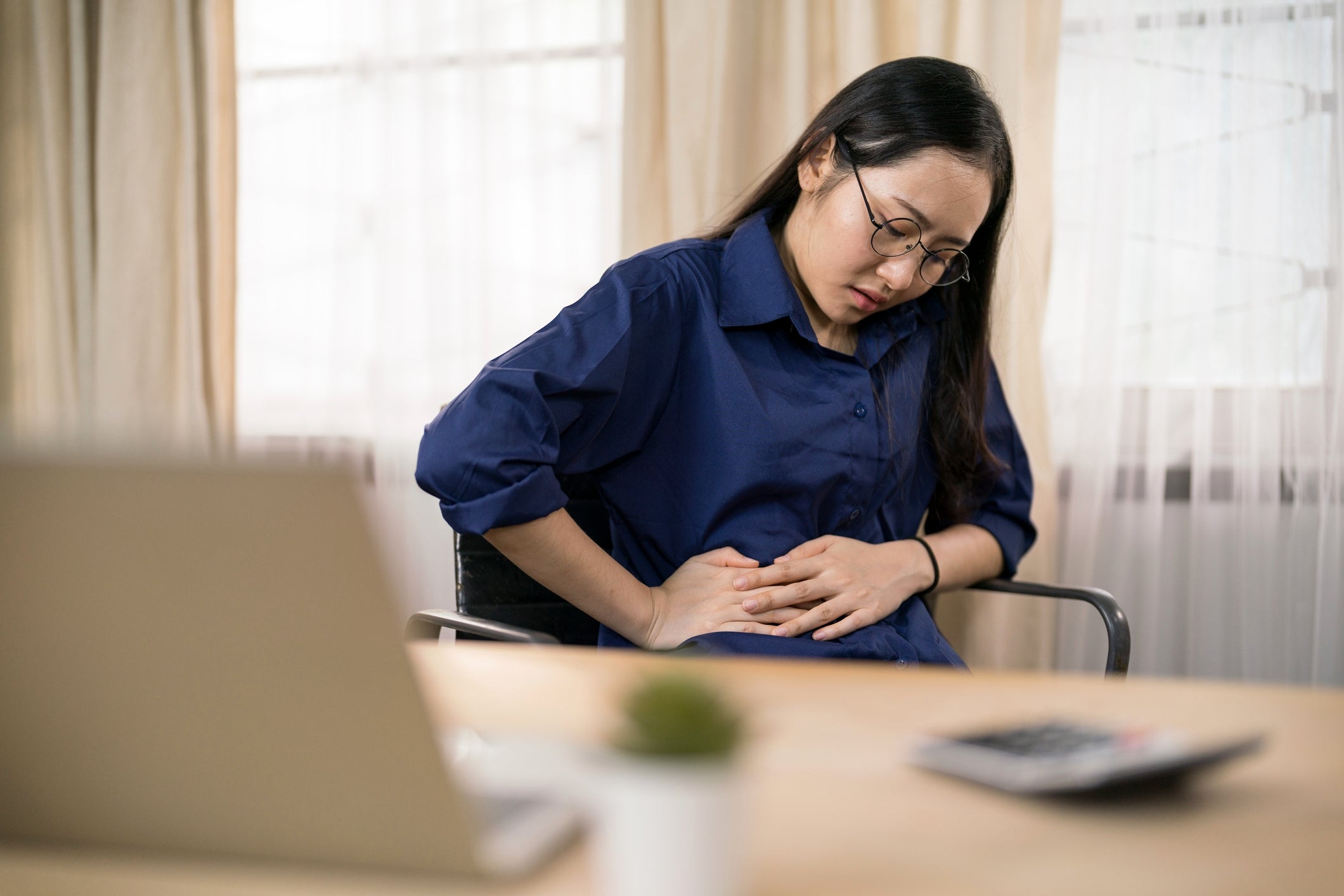 Person seated indoors, holding their stomach, looking unwell, near a laptop
