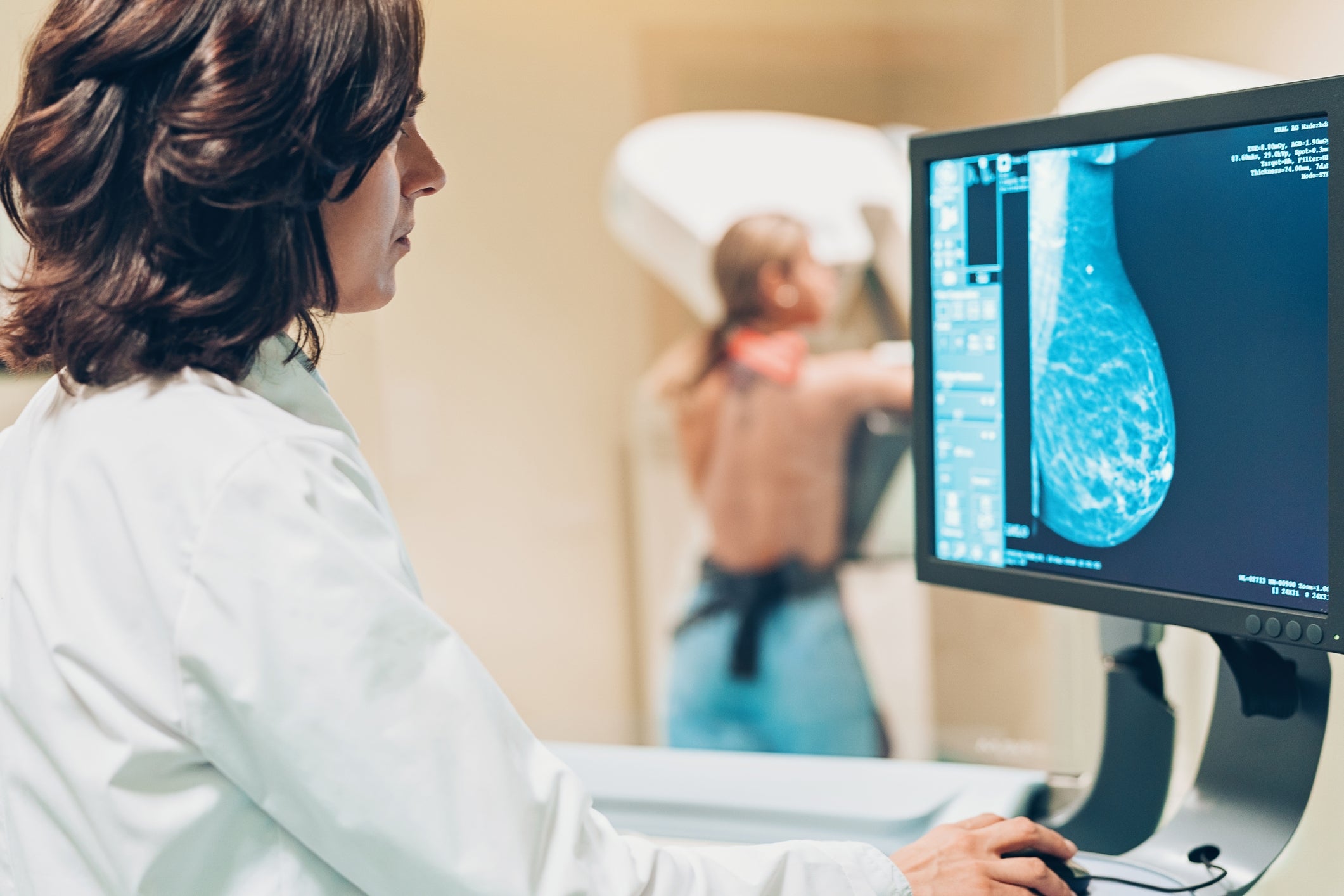 A technician examines a mammogram on a computer screen while a patient receives a breast scan in the background