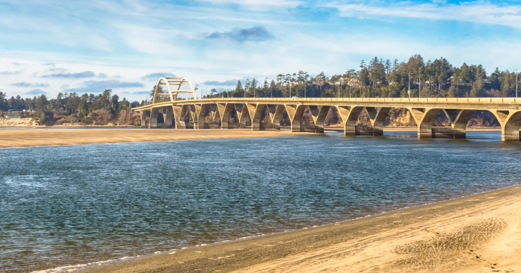 Scenic view of the Alsea Bay Bridge in Waldport, Oregon, spanning a river, surrounded by trees and a sandy shore under a blue sky.