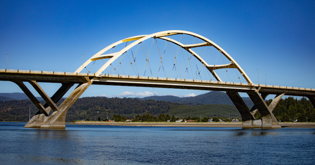 The Alsea Bay Bridge in Waldport, Oregon, with an arched design, spans across a river, surrounded by green hills and a clear blue sky.