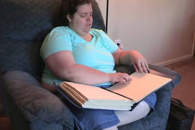 An individual is seated on a cushioned chair, immersed in reading a large, open book resting on their lap. The setting appears to be indoors, with a beige wall visible in the background.