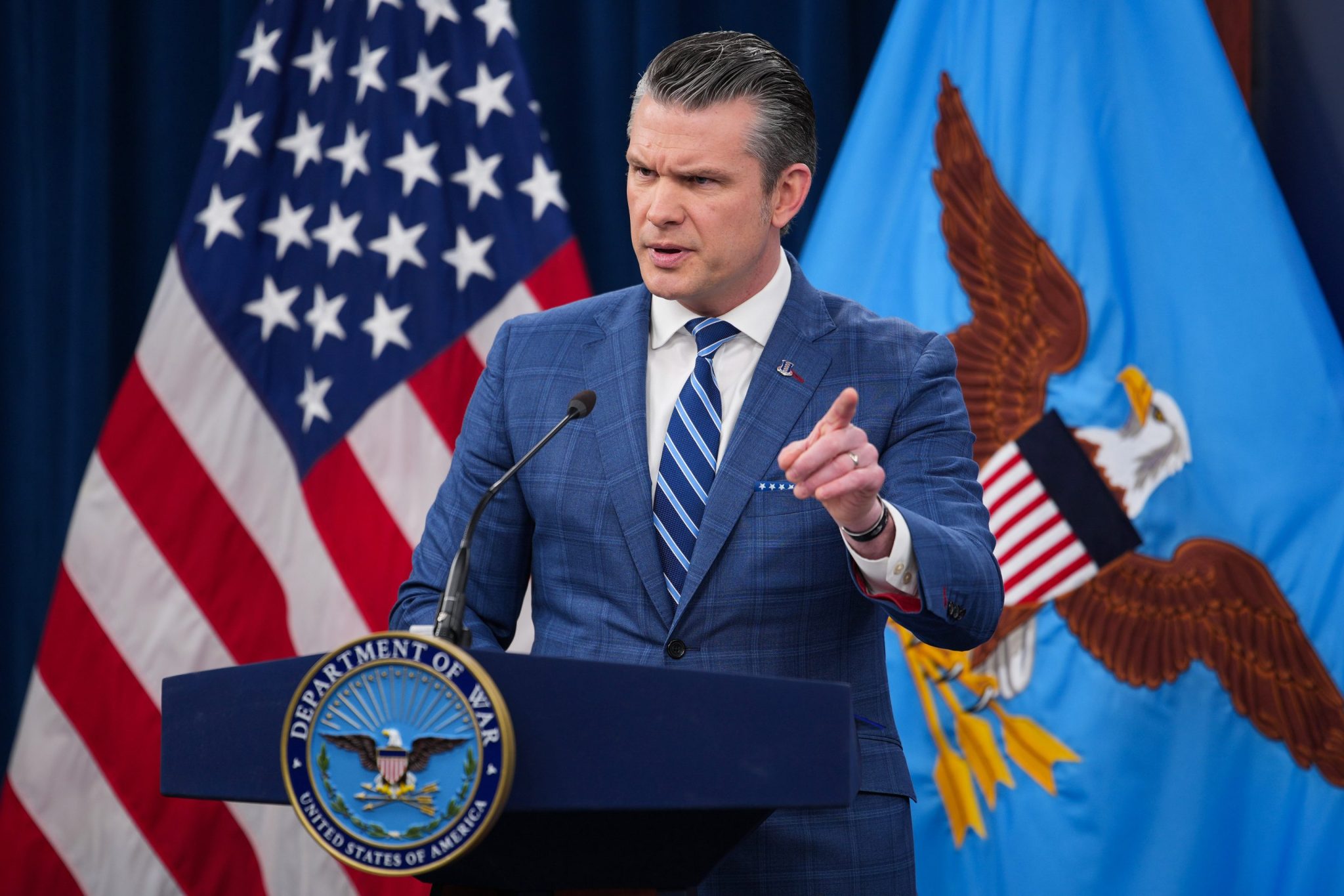 Pete Hegseth stands behind a podium, pointing with one hand and furrowing his brow.