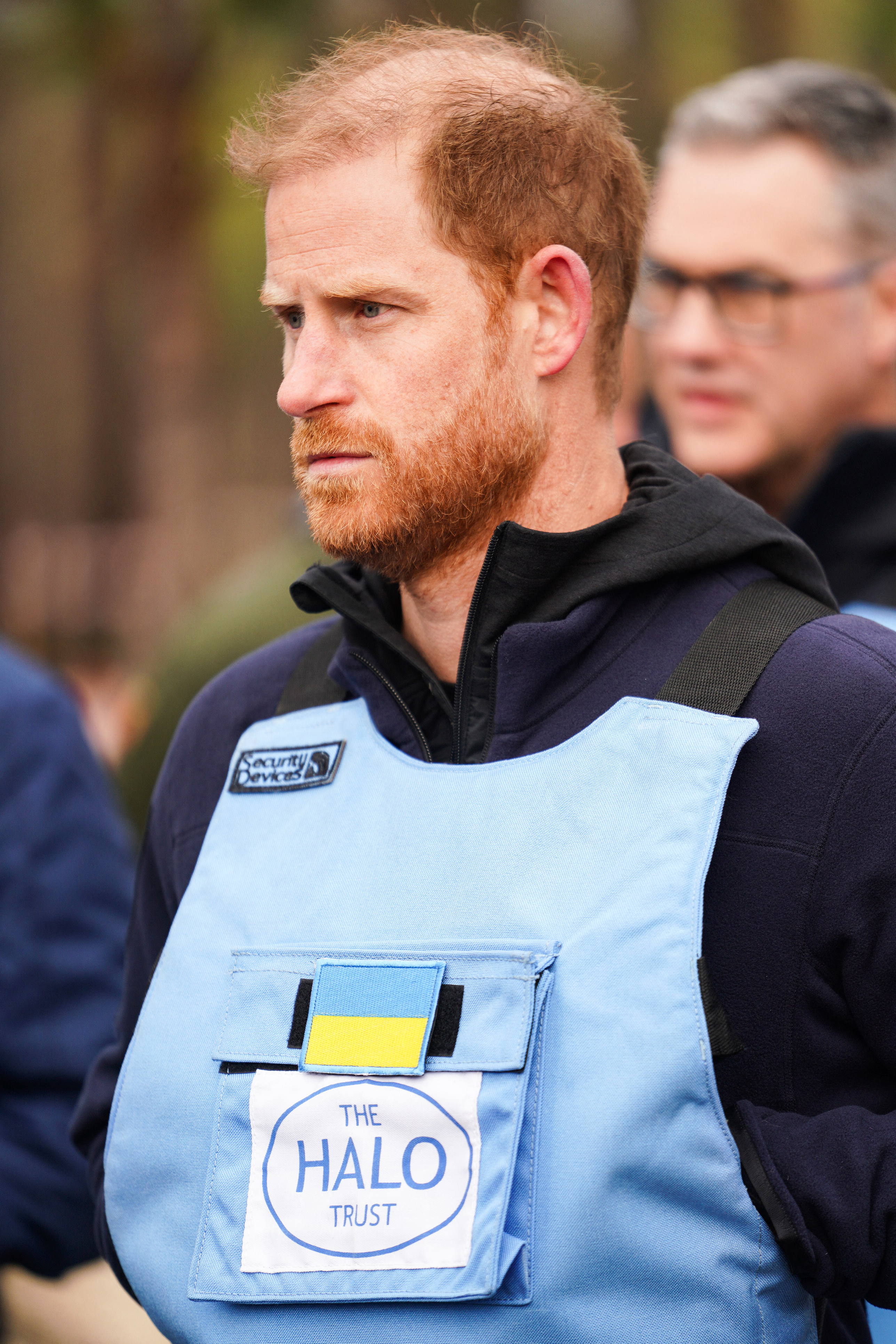 Prince Harry wearing a light blue vest with a Ukrainian flag patch and "The HALO Trust" logo, observing demining efforts near Bucha.