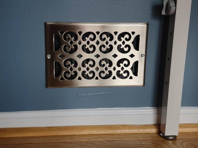 Decorative wall vent with an ornate, symmetrical metal grille, mounted near a light switch and above a hardwood floor