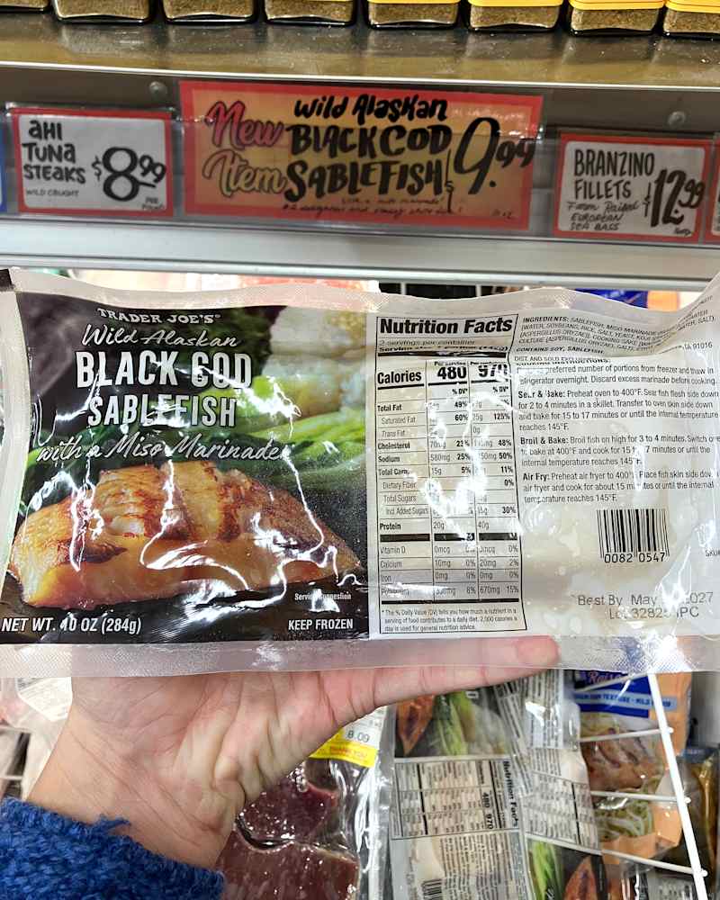 Package of Trader Joe's Wild Alaskan Black Cod Sablefish with a miso marinade, held in a hand, with price tags for other fish nearby.