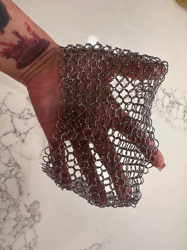 A hand wearing a chainmail glove holding a mesh bag, showing a tattoo. The scene is on a marble surface, related to shopping