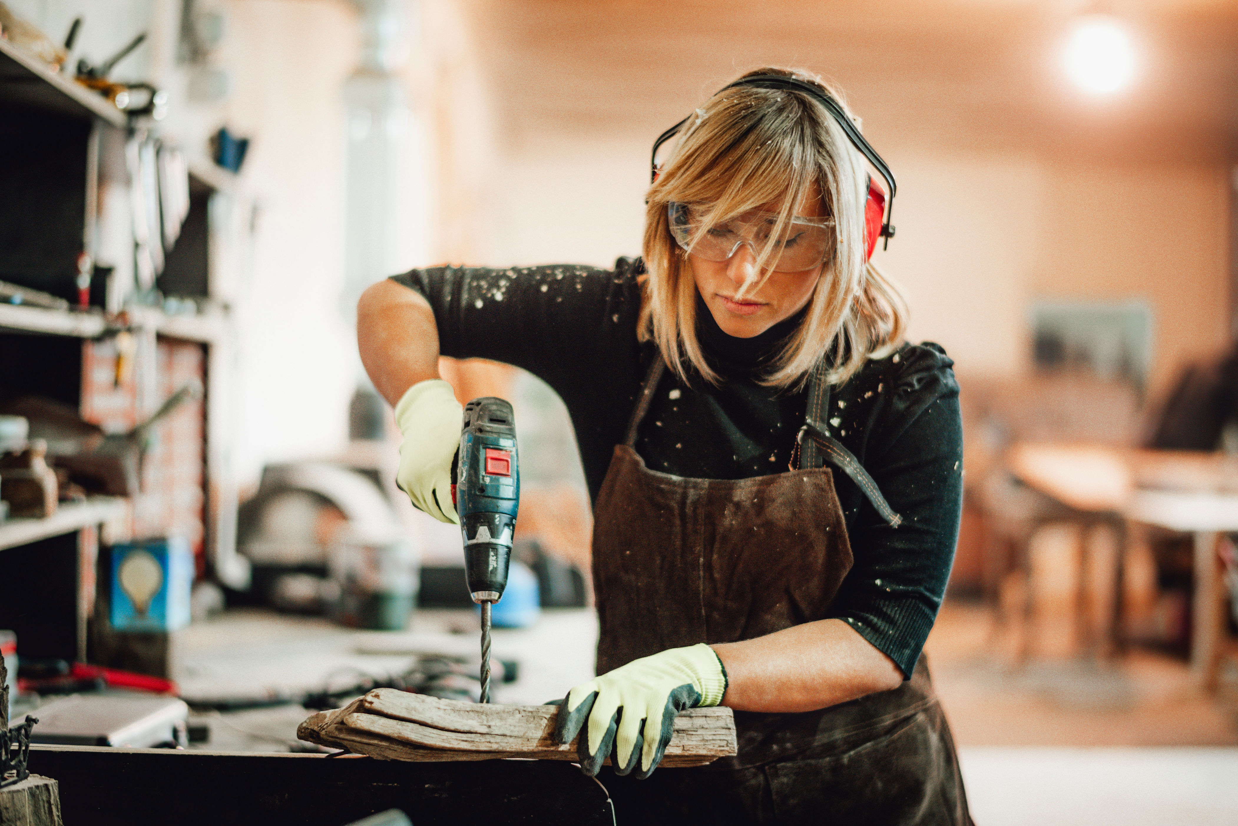 Person wearing safety gear uses a drill on a wooden board in a workshop, highlighting skilled craftsmanship and focus in a work environment