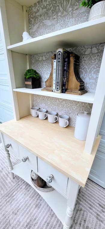 Wooden shelf with decor items, books, and white ceramic mugs. A decorative wallpaper background adds classic elegance