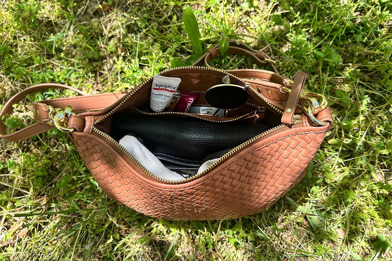 A woven crossbody bag on grass slightly open showing its contents like sunglasses and personal items
