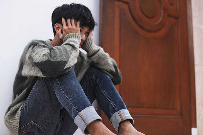 A frustrated teen, head in hands, sitting on the floor, contemplating moving out from family to sacrifice life.