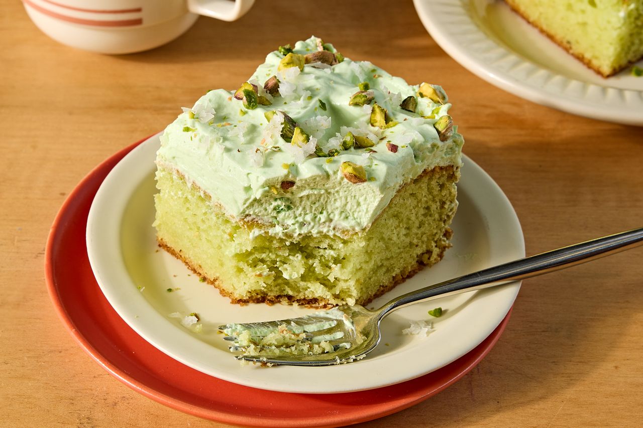 A slice of frosted cake topped with pistachios on a plate with a fork partial cake visible in the background