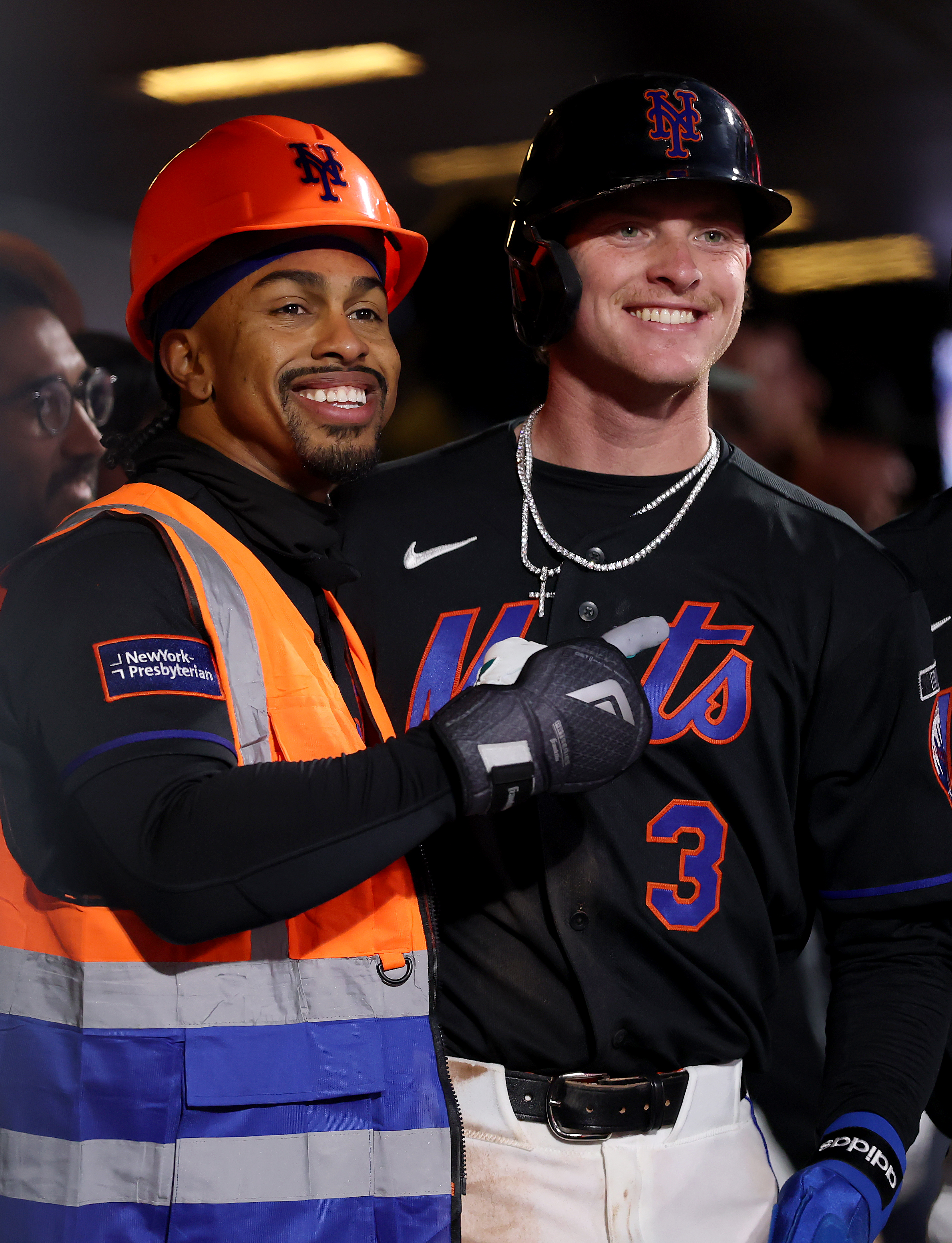 Francisco Lindor smiles in a black Mets uniform with an orange construction vest and helmet while standing next to Carson Benge