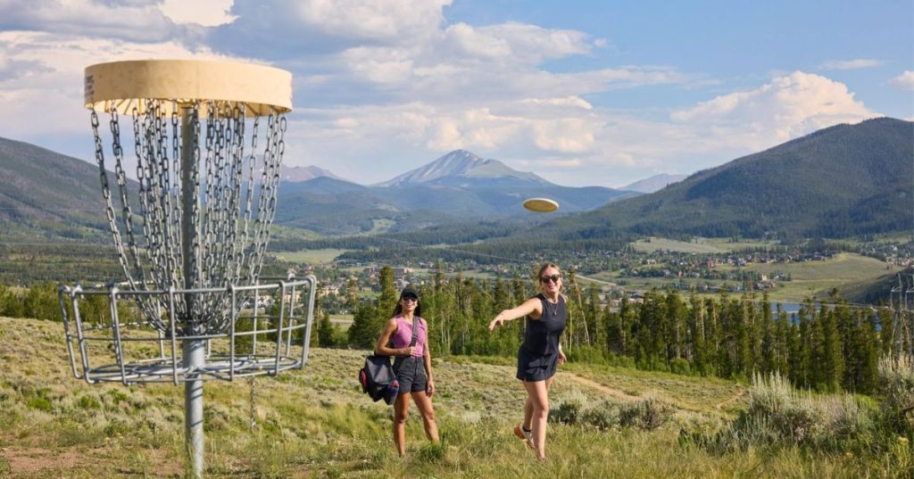 Two women enjoying a game of disc golf outdoors, with a disc basket in the foreground and a scenic mountain landscape in the background.