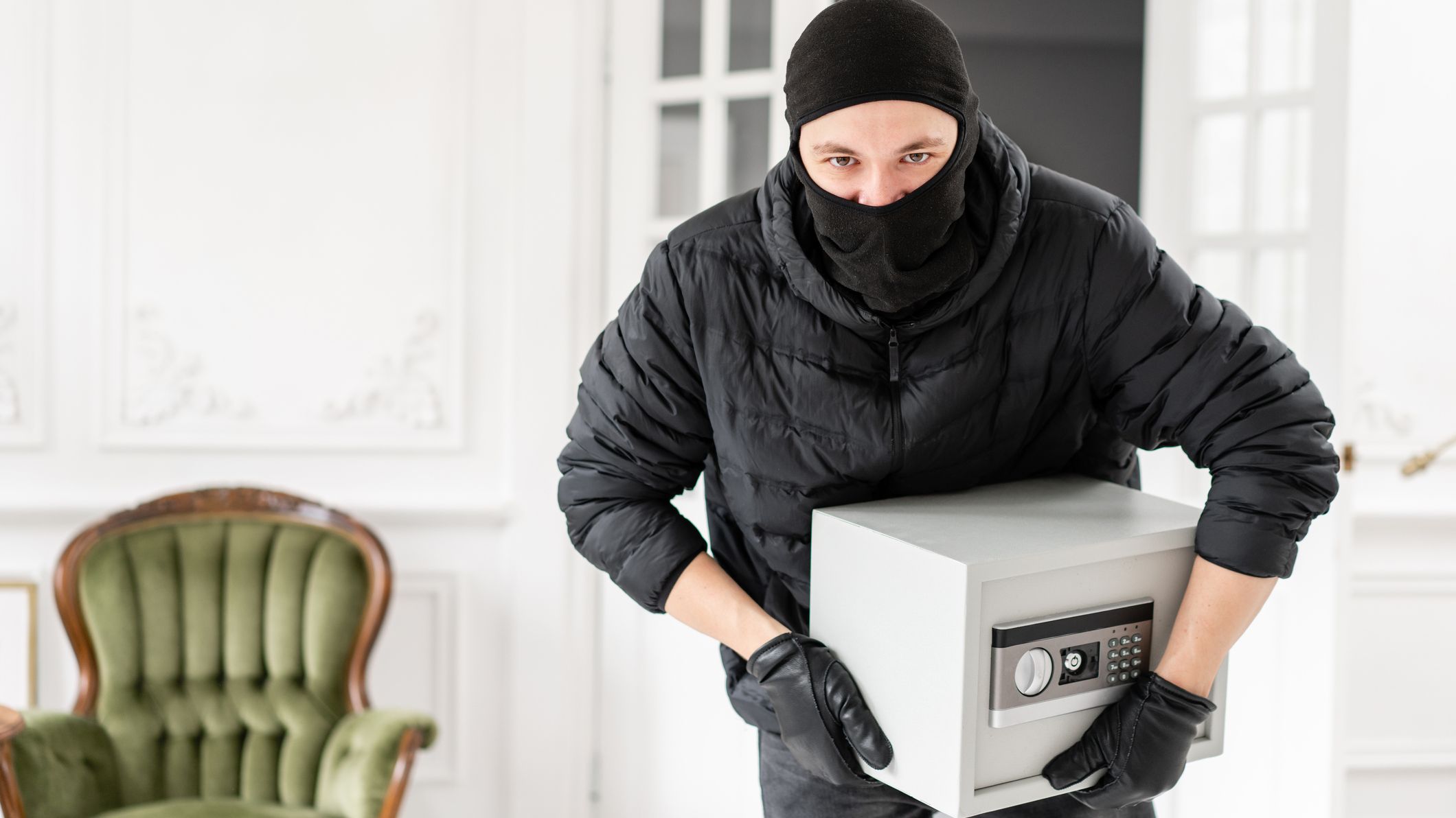 A thief wearing a mask and gloves carries a safe through a house.
